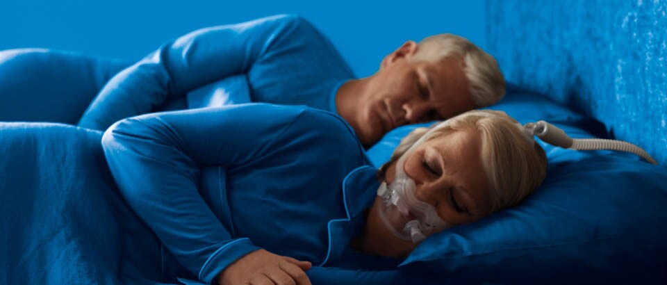 Vrouw in blauwe kleding die op haar zij ligt te slapen met een masker op en een man in blauwe kleding die op zijn zij ligt te slapen