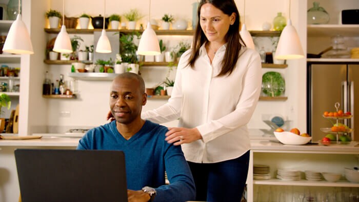 Foto van een man die in de keuken zit met zijn vrouw staand naast hem. Beiden kijken naar een laptopscherm