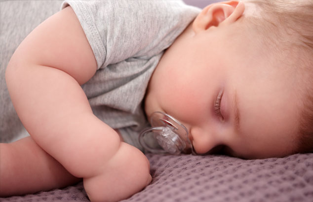 Een baby met een doorzichtige, ongevormde fopspeen in de mond slaapt op zijn zij op een paars dekentje.