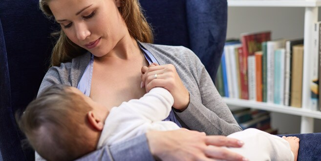 Een moeder zit achterover in een blauwe fluwelen stoel en kijkt neer op haar baby die borstvoeding krijgt in de madonnahouding.