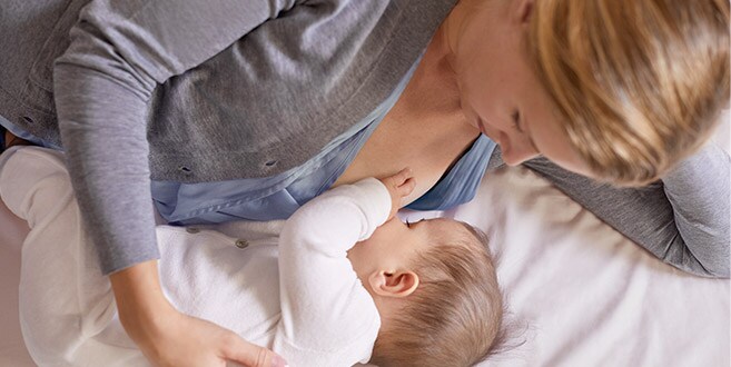 Een moeder ligt op haar linkerzij op een wit bed terwijl ze een baby borstvoeding geeft in liggende positie.