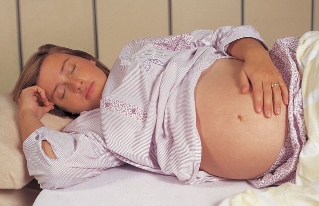 Een hoogzwangere vrouw slaapt op haar zij in bed met een hand op haar buik en de andere bij haar gezicht.
