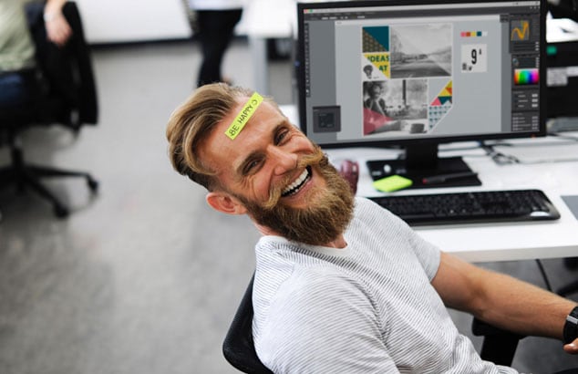 Blonde, jonge en leuk uitziende jongen met een grote baard die op een bureaustoel zit en naar de camera lacht