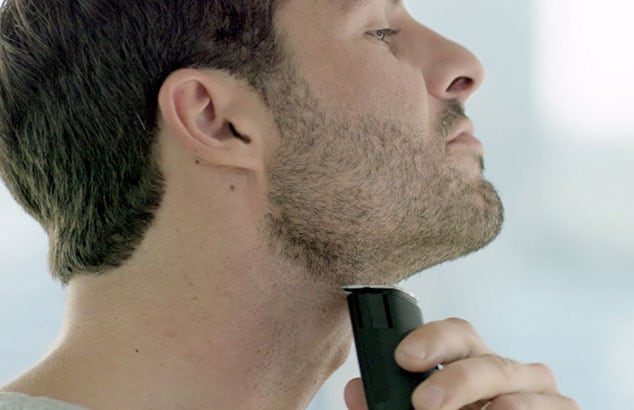 Zijprofiel van een man met korte baard die zijn hals scheert met een elektrisch scheerapparaat.