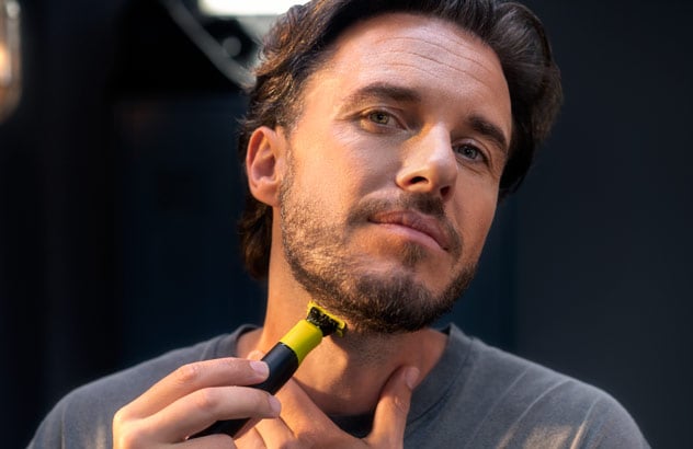 Close-up van een man die de rand van zijn donkere baard trimt met de Philips OneBlade voor een spiegel