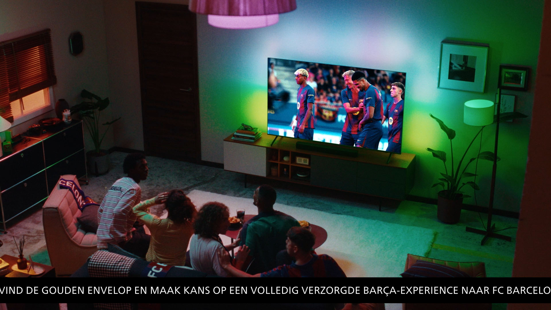 Een groep mensen kijkt naar een wedstrijd van FC Barcelona op een Philips Ambilight-tv, terwijl kleurrijke sfeerverlichting de woonkamer vult.